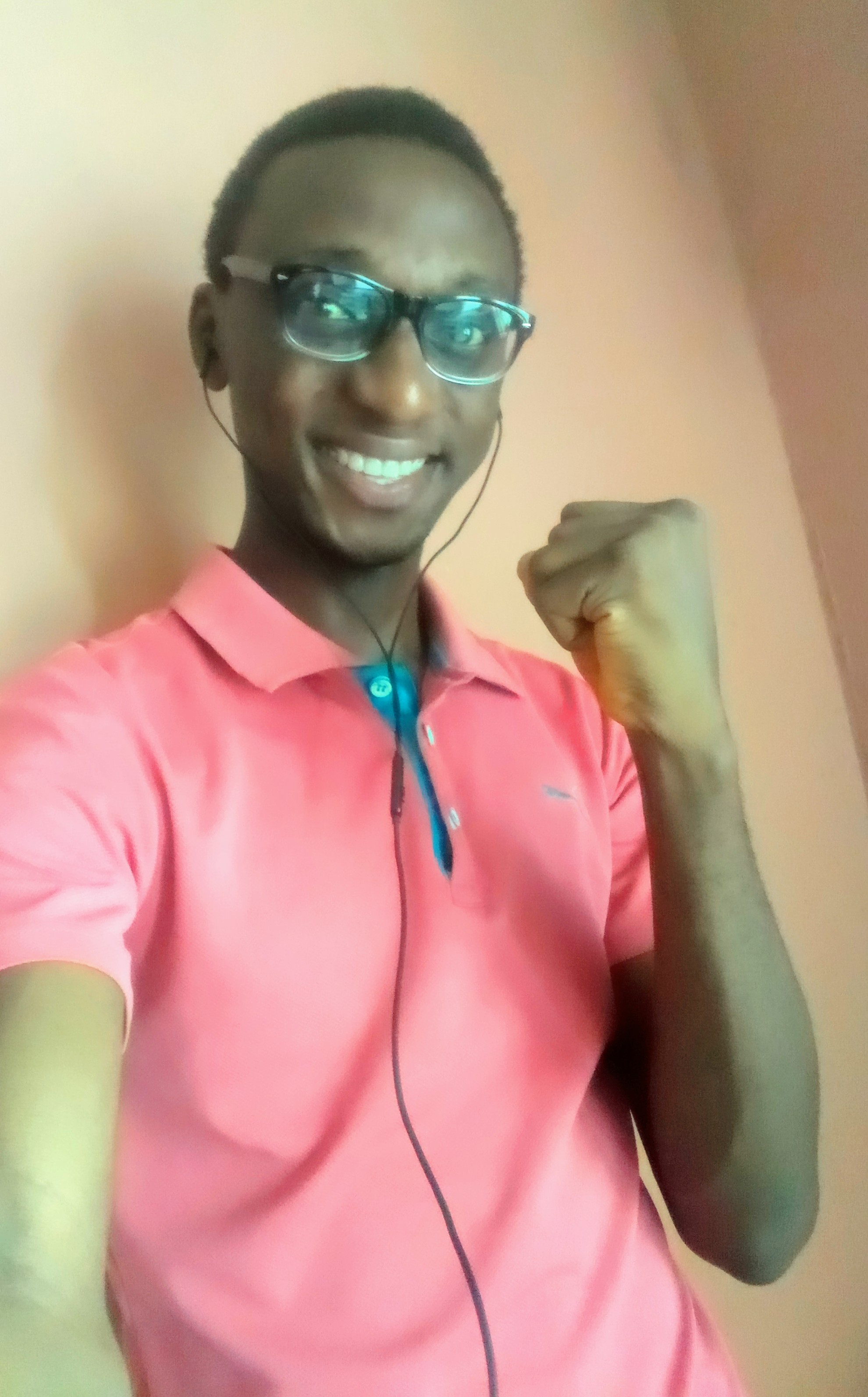Lewis smiling big time to the camera with a set forward optimistic fist, a pink t-shirt, a transparent eyeglass and black ear phone, looking very lively and excited.